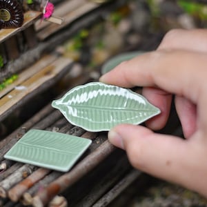 May include: Two small green ceramic plates shaped like leaves. The plates are on a wooden surface. One plate is being held by a hand.