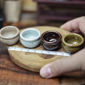 Puede incluir: Cuatro cuencos pequeños de cerámica en varios colores: beige, azul claro, marrón oscuro y verde oliva. Los cuencos están dispuestos sobre un disco de madera con una cinta métrica. Los cuencos son en miniatura y probablemente decorativos.