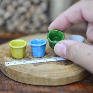 Puede incluir: Pequeños tiestos de cerámica en amarillo, azul, verde y gris, expuestos sobre una superficie de madera. Una cinta métrica se encuentra junto a los tiestos. Una mano sostiene el tiesto verde. Estos pequeños tiestos son probablemente decorativos.