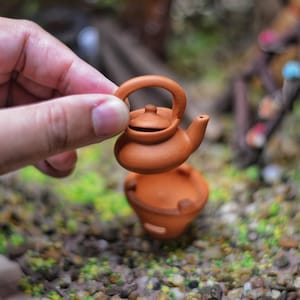 May include: A miniature terracotta teapot with a handle and spout is held by a hand. Below it, a matching terracotta pot is visible. The objects are set against a backdrop of small stones and greenery, suggesting a garden or miniature scene.