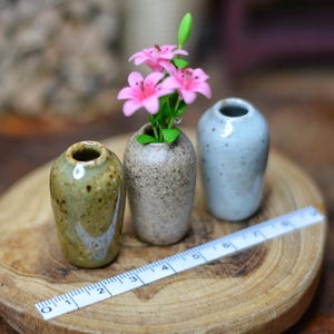 May include: Three miniature ceramic vases in green, beige, and blue-grey sit on a wooden surface. The centre vase holds pink flowers. A ruler is placed next to the vases. The vases are approximately 4 cm tall.