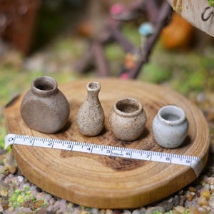 Może przedstawiać: Cztery miniaturowe ceramiczne wazony o różnych kształtach i kolorach stoją na drewnianym dysku z miarką. Wazony są w odcieniach brązu i bieli. Dysk leży na podłożu z małych kamieni i zieleni. Wazony mają wysokość od około 2,5 do 5 cm.