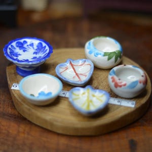 May include: A collection of small, hand-painted ceramic bowls and dishes in various shapes and designs. The bowls are white with blue, red, and green floral patterns. They are arranged on a round wooden tray, with a measuring tape.