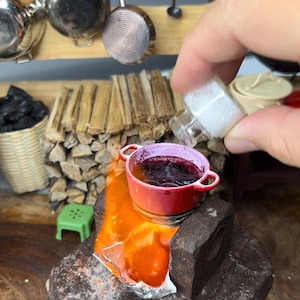Könnte beinhalten: Ein kleiner roter Topf mit einer violetten Flüssigkeit darin wird über einem kleinen Herd mit einer Flamme erhitzt. Eine Hand hält eine kleine Glasflasche mit weißen Kristallen über dem Topf.