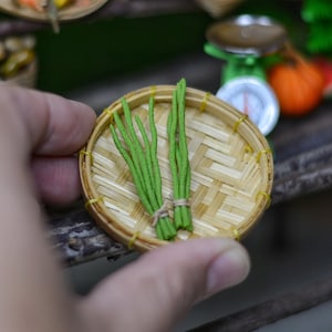 May include: A miniature woven bamboo basket holds a bundle of green chives tied with twine. The basket is round with a woven pattern and a yellow border. The chives are bright green.