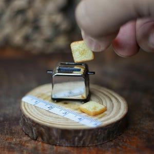 Op de afbeelding: Een miniatuur chromen broodrooster met een sneetje toast dat erin wordt geplaatst. Een ander stuk toast ligt naast de broodrooster op een houten schijf. Een meetlint is ook aanwezig.