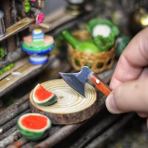 May include: A miniature wooden cutting board with a small watermelon slice and a whole watermelon on it. A small hand holds a miniature axe over the watermelon slice.