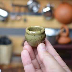 May include: A small, handmade ceramic cup with a speckled, olive-green glaze. The cup is held in a hand, showcasing its rounded shape and the texture of the glaze. The background is blurred, suggesting a kitchen setting.