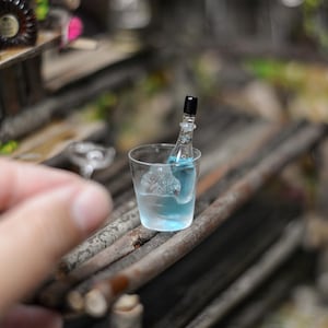 May include: A miniature glass with a blue liquid and a miniature bottle with a black cap inside. The glass is sitting on a wooden surface.
