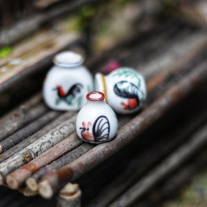 Könnte beinhalten: Drei kleine, weiße Keramikgläser mit Hahnmotiven in Rot, Schwarz und Grün. Die Gläser sind auf einer rustikalen Holzoberfläche aus kleinen Ästen angeordnet. Die Gläser haben einen roten Rand.