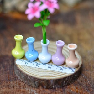 May include: Miniature ceramic vases in yellow, blue, white, lavender, and peach colors are arranged on a wooden slice. Pink flowers with green stems are in one vase. A ruler is placed in front of the vases.