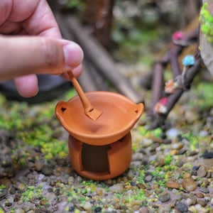 Op de afbeelding: Een kleine kleipot met een klein houten lepel erin. De pot is bruin en heeft een ronde vorm. De lepel rust op de rand van de pot.