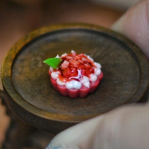 Puede incluir: Miniatura de tarta de fresas sobre un plato de madera pequeño. La tarta tiene una base de masa roja, relleno de crema blanca y una cobertura de fresas en dados y una hoja verde. El plato lo sostiene una persona.