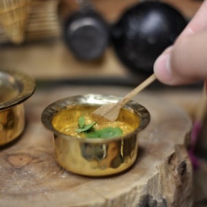 Puede incluir: Un pequeño cuenco dorado lleno de un plato de curry amarillo cubierto con hierbas verdes frescas. El cuenco está sobre una superficie de madera con una cuchara de madera cerca.