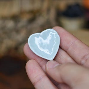 Puede incluir: Un pequeño plato de cerámica en forma de corazón de color azul pálido. El plato tiene un acabado brillante y está sostenido en la mano de una persona. El plato en forma de corazón es un objeto decorativo.