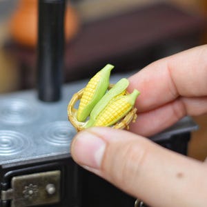 Könnte beinhalten: Miniatur Maiskolben in einem Weidenkorb. Der Mais hat gelbe Körner und grüne Blätter. Der Korb wird in der Nähe eines Miniaturherds gehalten, was auf ein Küchen- oder Lebensmittelthema hindeutet.