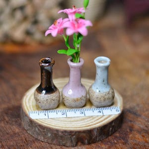 Puede incluir: Tres pequeños jarrones de cerámica en tonos marrón, rosa y azul, cada uno con una base moteada, se exhiben sobre una rodaja de madera. El jarrón rosa contiene pequeñas flores rosas. Una regla está colocada frente a los jarrones.