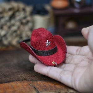 Könnte beinhalten: Miniatur-Cowboyhut in Rot mit braunem Band und silbernem Stern. Der Hut hat eine kleine Krempe und ist als Anhänger oder Anhänger gedacht. Der Hut wird in einer Hand gehalten.
