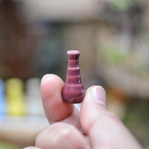 Könnte beinhalten: Eine kleine, handgefertigte Miniaturvase in Burgunder- und Weißtönen. Die Vase hat einen schlanken Hals und einen bauchigen Körper mit horizontalen Streifen. Die Vase wird zwischen zwei Fingern gehalten, was ihre zarte Größe und ihr aufwendiges Design zeigt.