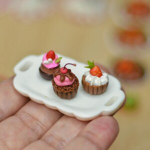 Puede incluir: Pequeños postres en una bandeja blanca. Tres cupcakes diminutos están decorados con glaseado rosa, una cereza y una fresa. La bandeja tiene un borde festoneado y dos asas. Los postres son marrones, rosas y blancos.