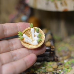 Pode incluir: Uma pequena cesta de vime cheia de cenouras brancas em miniatura com pontas verdes. A cesta está sendo segurada na mão de uma pessoa.