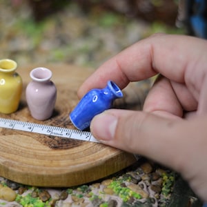 Może przedstawiać: Miniaturowe ceramiczne wazony w kolorze żółtym, różowym i niebieskim są ułożone na drewnianym plastrze z miarką. Niebieski wazon jest trzymany w dłoni. Wazony mają około 2,5 cm wysokości, idealne do domku dla lalek.