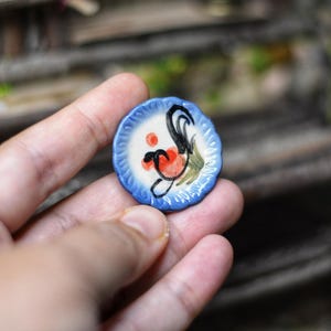 May include: A small, round, hand-painted ceramic plate with a blue rim and a white center. The plate features a rooster design in black, red, and green. The plate is held in a person's hand.