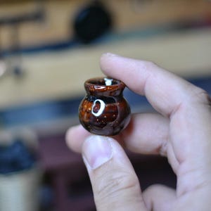 May include: A small, brown ceramic pot with a glossy finish. The miniature pot has a rounded body and a small opening at the top. The pot is held between fingers, showcasing its size and detail. The pot is likely a decorative item.