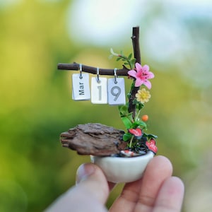 Puede incluir: Calendario decorativo en miniatura con la fecha "Mar 19" mostrada en pequeñas etiquetas blancas. El calendario está adornado con flores rosas y amarillas, bayas rojas y un pequeño trozo de madera, todo en una maceta de cerámica blanca.