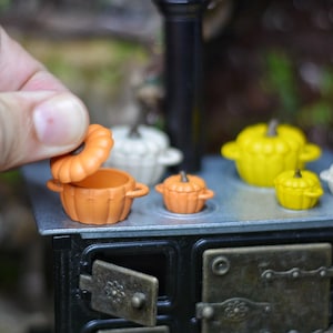 May include: Miniature orange pumpkin-shaped pot with a lid being opened, placed on a small black stove. Other miniature pots in white and yellow are also on the stove. The scene evokes a whimsical, autumnal theme.
