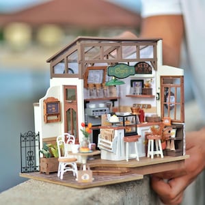 May include: Miniature cafe model with a detailed interior, including a counter, tables, and chairs. The model features a sign that says "Cafe" and a small black metal fence. The cafe is made of wood and has a glass roof.