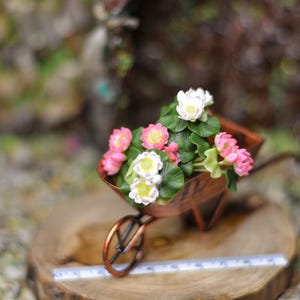 以下が含まれることがあります： 緑の葉とピンクと白の花で満たされたミニチュアの銅製手押し車。手押し車は木の輪切りに置かれ、前に定規があります。花の中心は黄色です。