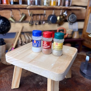 Könnte beinhalten: Drei kleine Gewürzgläser aus Glas mit Schraubdeckeln. Die Gläser sind mit "Salz", "Chili" und "Weißer Pfeffer" beschriftet. Die Gläser stehen auf einem kleinen Holzhocker.