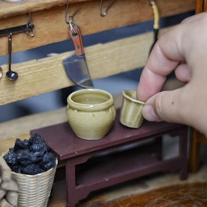 May include: Miniature kitchen scene with a wooden shelf holding small cooking utensils and a knife. A small, round, light-colored pot and a small cup sit on a miniature table. A hand is holding the cup. A basket of coal and a stack of wood are in the foreground.