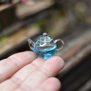 Puede incluir: Una tetera de vidrio en miniatura con un líquido azul en su interior. La tetera está sentada en la mano de una persona.