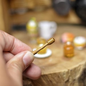 Può includere: Un piccolo cucchiaino di miele in legno, tenuto in mano. Il cucchiaino ha una testa scanalata e un manico sottile. Altri oggetti alimentari in miniatura sono sullo sfondo, suggerendo una casa delle bambole o un progetto artigianale.