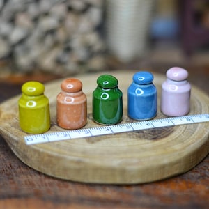 May include: Five miniature ceramic jars in yellow, orange, green, blue, and pink sit on a wooden disc. Each jar has a rounded lid. A white ruler with black markings, showing measurements in centimetres, is placed in front of the jars. The background is blurred.