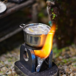 Pode incluir: Um fogão em miniatura com uma panela pequena em cima. O fogão está aceso e tem uma chama saindo do lado. A panela está cheia de um líquido branco.