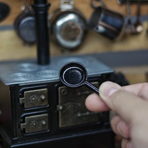 Könnte beinhalten: Miniatur-schwarze Bratpfanne vor einem kleinen schwarzen und silbernen Ofen. Der Ofen hat eine Metallplatte mit vier Kochfeldern und zwei kleinen Ofentüren. Küche-Themen-Miniaturdekoration.