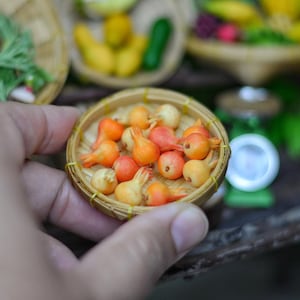 Puede incluir: Una pequeña cesta redonda de mimbre llena de pequeñas frutas naranjas y amarillas en miniatura. La fruta tiene forma bulbosa con un pequeño tallo. En el fondo hay otras cestas en miniatura con productos, junto con una pequeña balanza y una calabaza.