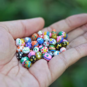 May include: A handful of colorful, decorative beads, each uniquely painted with patterns like polka dots, stripes, and floral designs. The beads are in various colors, including blue, pink, yellow, and green. The beads are small and round, likely for crafting or jewelry making.