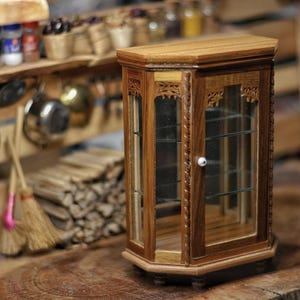 Op de afbeelding: Een miniatuur houten vitrinekast met glazen deuren en planken. De kast is gemaakt van donker hout met ingewikkelde gravures en een kleine witte knop. Hij staat op een houten oppervlak, met een rustieke keukenscène op de achtergrond.