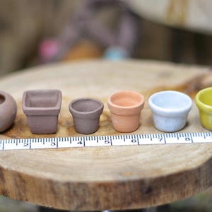 Może przedstawiać: Kolekcja sześciu miniaturowych ceramicznych doniczek w różnych kształtach i kolorach, w tym brązowym, brzoskwiniowym, białym i żółtym. Doniczki są ułożone na drewnianej powierzchni z linijką dla skali, co sugeruje, że są przeznaczone do małych roślin lub rękodzieła.