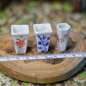 Può includere: Tre vasi in miniatura in ceramica bianca con disegni floreali rossi e blu. I vasi sono quadrati ed esagonali, appoggiati su una fetta di legno con un metro a nastro. I vasi sono alti circa 2,5-4 cm.