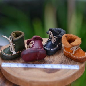 Könnte beinhalten: Vier Miniatur-Lederstiefel in Grün, Burgunderrot, Schwarz und Orange, jeweils mit Schnürsenkeln. Die Stiefel sind auf einer Holzoberfläche angeordnet, mit einem Lineal davor. Die Stiefel sind ein Bastelartikel.