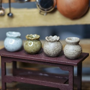 May include: Four miniature ceramic vases in varying shades of blue, green, and brown sit on a small, dark red table. The vases have unique shapes and textures, with a rustic, handmade appearance. Kitchen utensils hang in the background.
