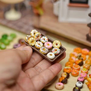 May include: Miniature donuts in a brown tray, held in a hand. The donuts are decorated with colorful sprinkles and frosting. Other miniature pastries are arranged on a wooden surface, including croissants and glazed donuts. A miniature bakery scene is in the background.
