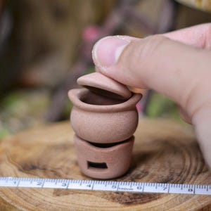Può includere: Una piccola pentola in ceramica beige a due pezzi con coperchio. La pentola è su una superficie di legno. Un metro a nastro è posto accanto alla pentola, che mostra le misure in centimetri.