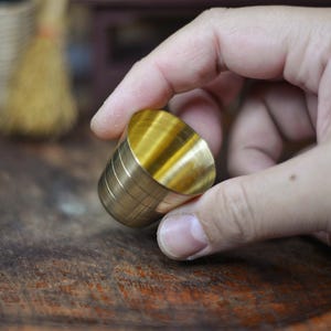 May include: A cylindrical brass object with a golden hue, held in a hand. The object has a slightly tapered shape and horizontal grooves around its body. The background is a blurred wooden surface.