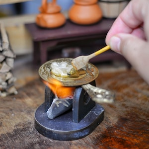 Puede incluir: Escena de cocina en miniatura con una pequeña estufa, un plato con comida y una pequeña cuchara de madera. La estufa tiene una llama y el plato se está removiendo. Otros objetos en miniatura están en el fondo.